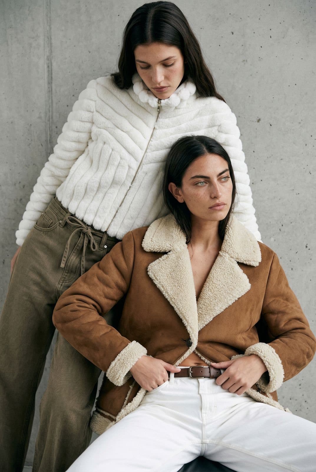 Custom developed outerwear: a white textured jacket and a brown shearling coat, on two models against a concrete wall.